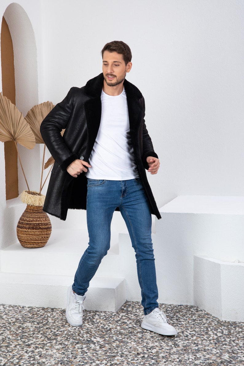 Puede incluir: Un hombre con un abrigo de piel de oveja negro, una camiseta blanca y pantalones vaqueros azules. Est&aacute; de pie en una plataforma blanca con un fondo blanco.