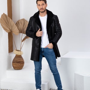 Puede incluir: Una chaqueta de piel de oveja negra con una camiseta blanca y unos vaqueros azules. La persona est&aacute; de pie frente a una pared blanca con una planta decorativa.