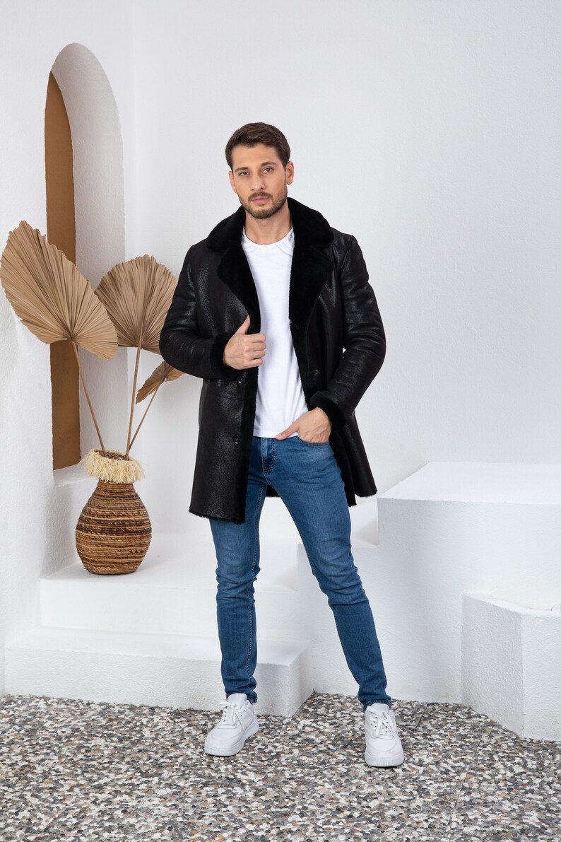 Puede incluir: Una chaqueta de piel de oveja negra con una camiseta blanca y unos vaqueros azules. La persona est&aacute; de pie frente a una pared blanca con una planta decorativa.