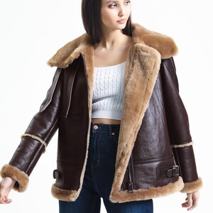 May include: A brown leather jacket with a shearling lining and trim. The jacket features a wide collar, a zip closure, and buckle details. The sleeves have shearling cuffs. The jacket is worn over a white top and blue jeans.