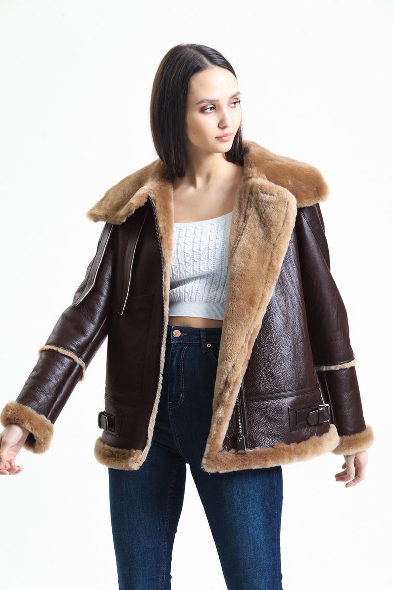 May include: A brown leather jacket with a shearling lining and trim. The jacket features a wide collar, a zip closure, and buckle details. The sleeves have shearling cuffs. The jacket is worn over a white top and blue jeans.