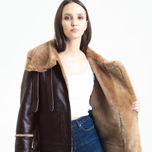 May include: A brown leather jacket with a shearling lining and collar. The jacket has a classic aviator style with a zip closure, buckle details, and shearling trim on the cuffs and hem. The jacket is worn open, revealing the shearling interior.
