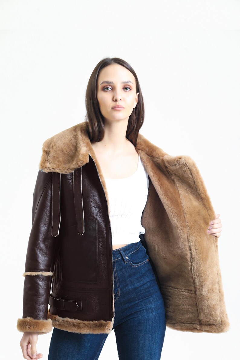 May include: A brown leather jacket with a shearling lining and collar. The jacket has a classic aviator style with a zip closure, buckle details, and shearling trim on the cuffs and hem. The jacket is worn open, revealing the shearling interior.