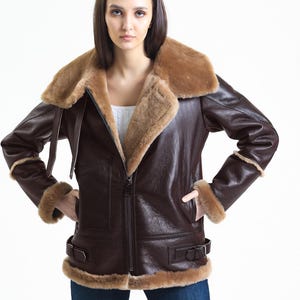 May include: A brown leather jacket with a shearling collar and trim. The jacket has a zip-up front, two pockets, and buckle details at the waist. The jacket is worn with blue jeans.