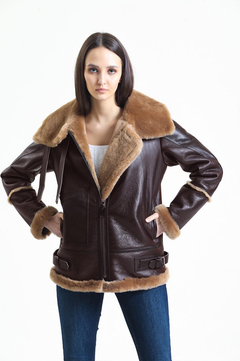 May include: A brown leather jacket with a shearling collar and trim. The jacket has a zip-up front, two pockets, and buckle details at the waist. The jacket is worn with blue jeans.