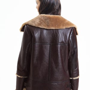 May include: A brown leather jacket with a tan shearling collar and trim. The jacket has a classic aviator style with a wide collar and a tailored fit. The back view shows the jacket's details, including the seams and the texture of the leather.