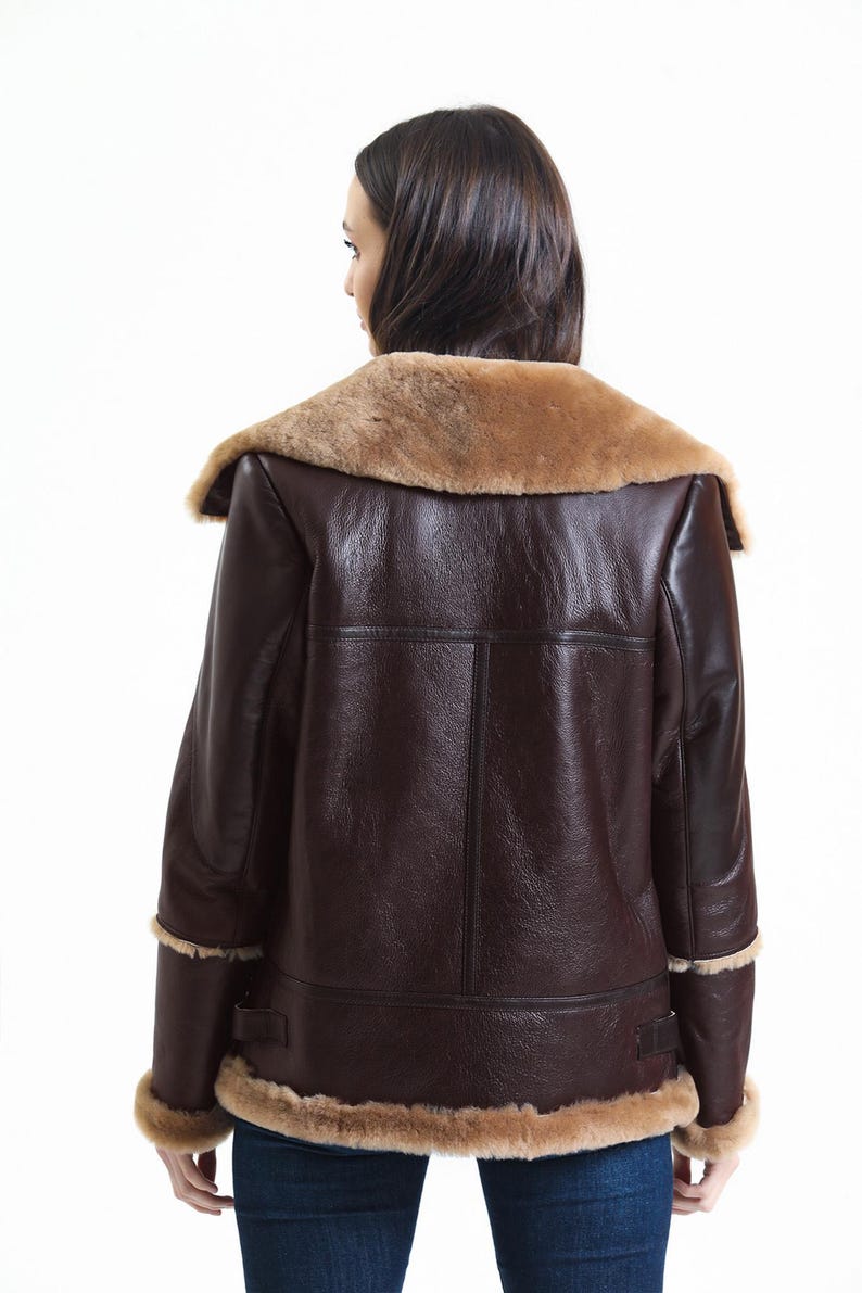 May include: A brown leather jacket with a tan shearling collar and trim. The jacket has a classic aviator style with a wide collar and a tailored fit. The back view shows the jacket's details, including the seams and the texture of the leather.