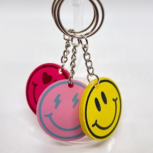May include: Three keychains with different smiley faces. The first keychain is pink with a blue lightning bolt for each eye and a blue smile. The second keychain is yellow with a black smile and two black eyes. The third keychain is red with a pink heart for an eye and a pink smile.