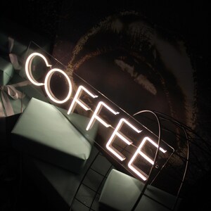 May include: White neon sign with the word "COFFEE" on a clear acrylic background. The sign is sitting on a black metal stand.