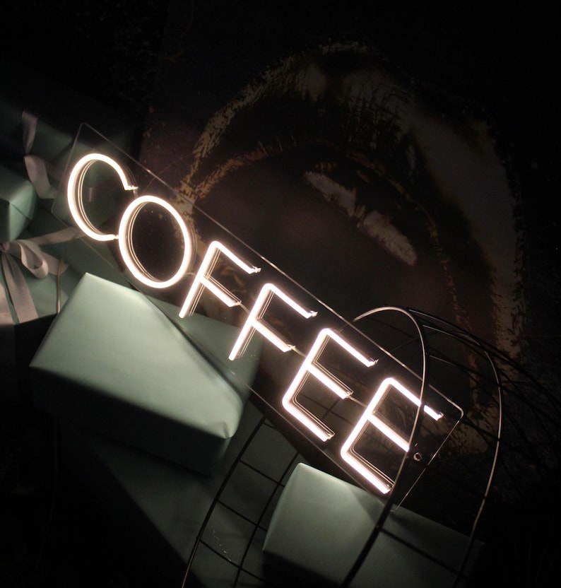 May include: White neon sign with the word "COFFEE" on a clear acrylic background. The sign is sitting on a black metal stand.