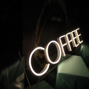 May include: White neon sign with the word "COFFEE" in a modern font. The sign is mounted on a clear acrylic panel and is illuminated from behind.