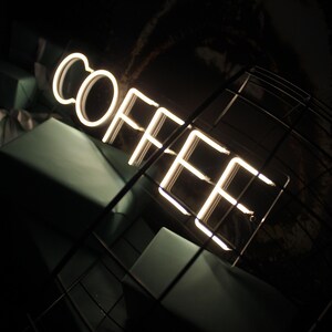May include: A white neon sign that reads "COFFEE" in a dark room with a black metal grid in the foreground.