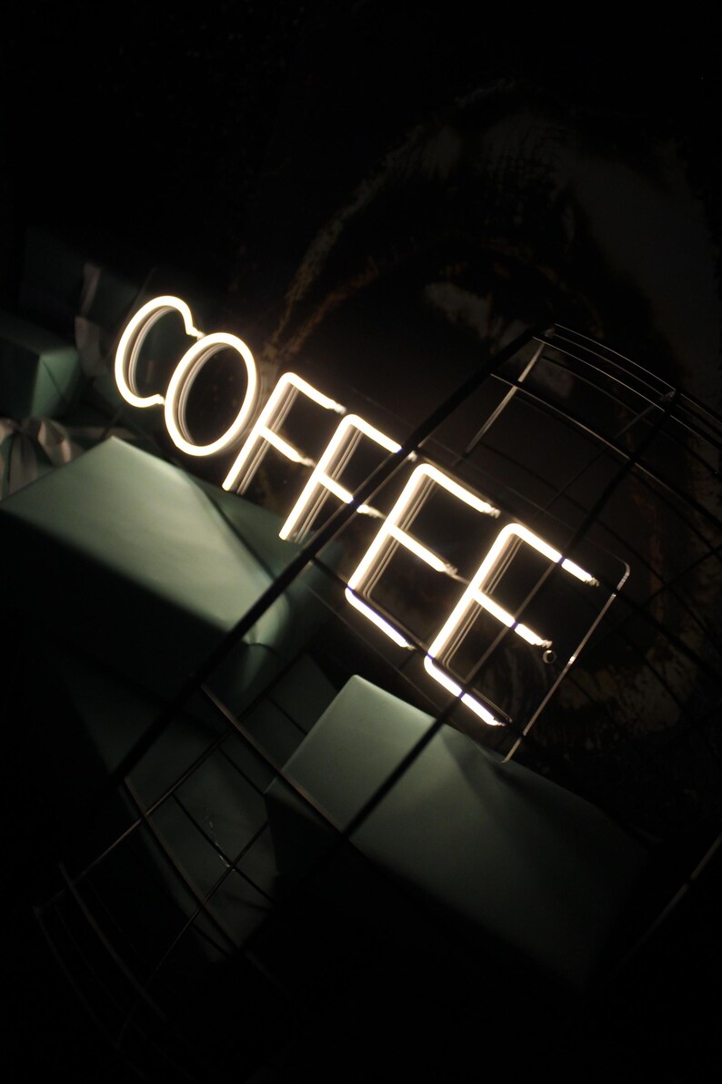 May include: A white neon sign that reads "COFFEE" in a dark room with a black metal grid in the foreground.