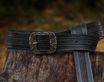May include: A black leather belt with a decorative brass buckle and intricate woven designs. The belt is wrapped around a tree stump.
