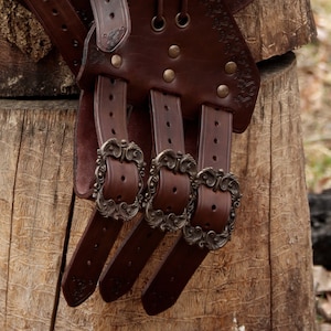 May include: A brown leather belt with multiple buckles and decorative metal accents. The belt is designed with a series of straps and buckles that can be adjusted to fit different sizes.