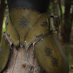 Könnte beinhalten: Ein grünes Leder-Rüstungsset mit komplizierten Designs und einer kleinen Tasche, die an den Gürtel befestigt ist. Die Rüstung wird von einem Mannequin getragen und vor einem hölzernen Hintergrund ausgestellt.