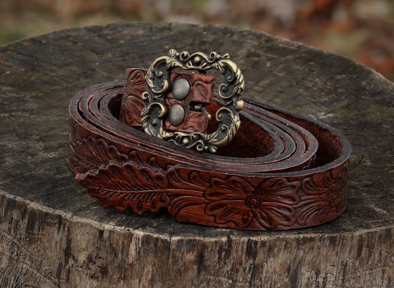 May include: A brown leather belt with a floral design and a decorative metal buckle. The belt is rolled up and the buckle is visible.