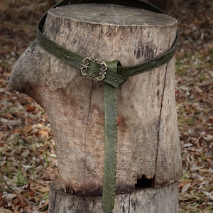 May include: A green leather belt with an ornate brass buckle, tied around a tree stump.