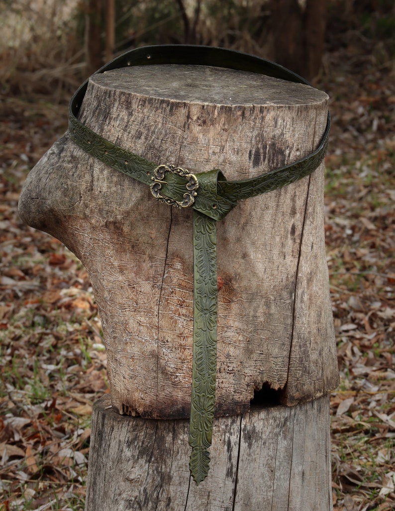 May include: A green leather belt with an ornate brass buckle, tied around a tree stump.