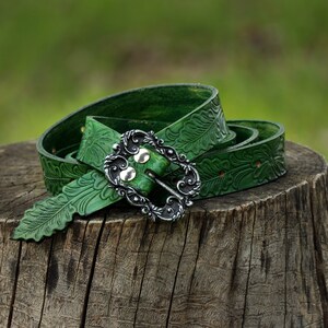 May include: A green leather belt with an ornate silver buckle and a leaf pattern. The belt is coiled on a tree stump.