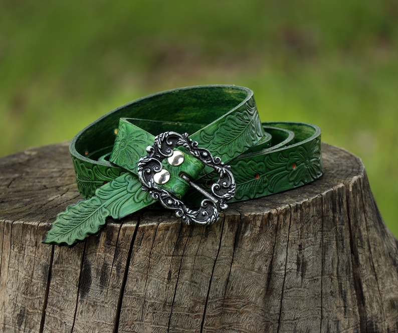 May include: A green leather belt with an ornate silver buckle and a leaf pattern. The belt is coiled on a tree stump.