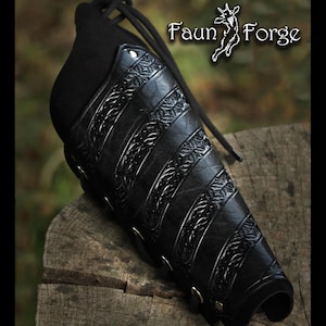 Puede incluir: Guardabrazos de cuero negro con diseños florales en relieve y ojales metálicos. El guardabrazos tiene un cordón negro para sujetarlo al brazo. Producto de Faun Forge.