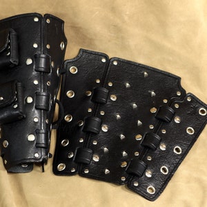 K&ouml;nnte beinhalten: Schwarze Leder-Armr&uuml;stung mit Metallnieten und Nieten. Die R&uuml;stung hat ein segmentiertes Design und beinhaltet eine Tasche mit einer Klappe.
