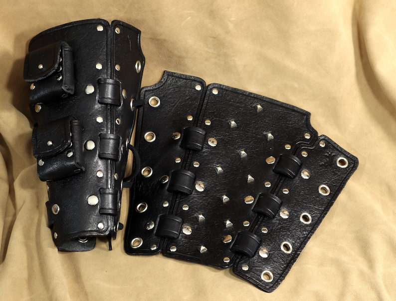 K&ouml;nnte beinhalten: Schwarze Leder-Armr&uuml;stung mit Metallnieten und Nieten. Die R&uuml;stung hat ein segmentiertes Design und beinhaltet eine Tasche mit einer Klappe.