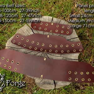 Puede incluir: Tres cinturones de cuero marrón con ojales y hebillas de latón. Los cinturones están dispuestos sobre una superficie de madera con hierba verde en el fondo. El texto "Hero Belt sizes:" se muestra encima de los cinturones, seguido de medidas en centímetros y pulgadas. El texto "Panel pratical lenght:" se muestra a la derecha de los cinturones, seguido de medidas en centímetros y pulgadas. El texto "Faun Forge" se muestra debajo de los cinturones.