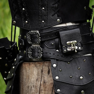 K&ouml;nnte beinhalten: Schwarzer Lederg&uuml;rtel mit silbernen Schnallen und einer kleinen Tasche. Der G&uuml;rtel ist mit silbernen Nieten verziert und hat eine dekorative Schnalle mit einem Metallverschluss.