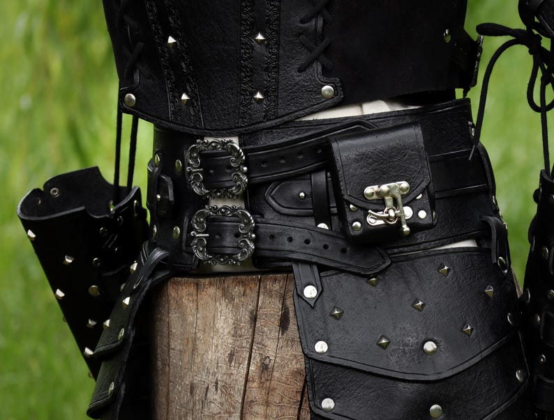K&ouml;nnte beinhalten: Schwarzer Lederg&uuml;rtel mit silbernen Schnallen und einer kleinen Tasche. Der G&uuml;rtel ist mit silbernen Nieten verziert und hat eine dekorative Schnalle mit einem Metallverschluss.