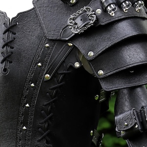 K&ouml;nnte beinhalten: Schwarze Leder-R&uuml;stung mit einer silbernen Schnalle und dekorativen Details. Die R&uuml;stung hat eine geschn&uuml;rte Vorderseite und eine Reihe von Riemen und Schnallen.