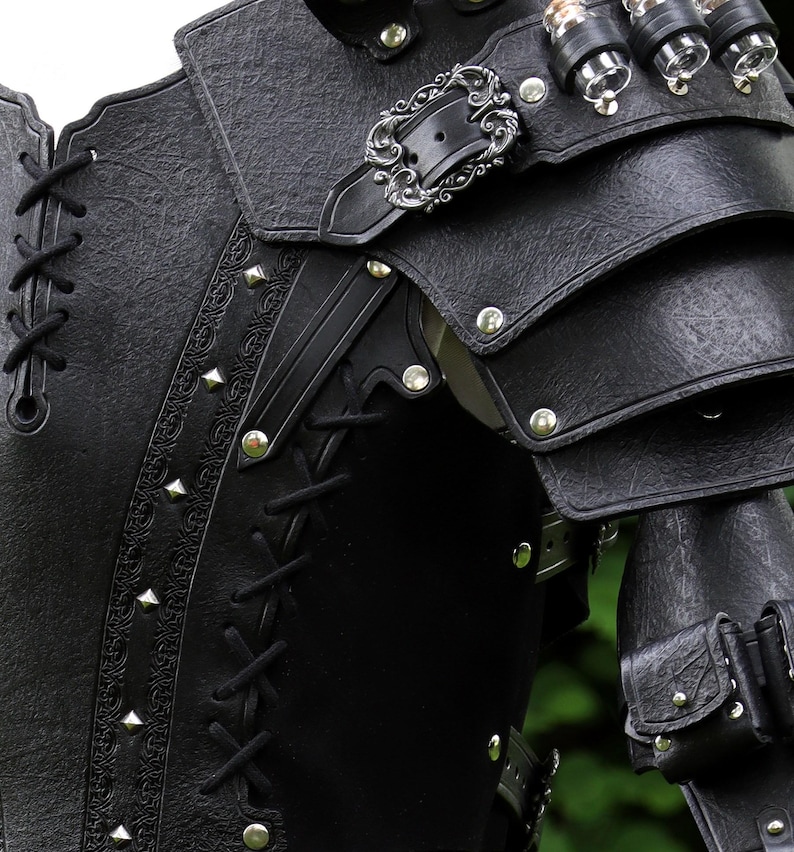 K&ouml;nnte beinhalten: Schwarze Leder-R&uuml;stung mit einer silbernen Schnalle und dekorativen Details. Die R&uuml;stung hat eine geschn&uuml;rte Vorderseite und eine Reihe von Riemen und Schnallen.