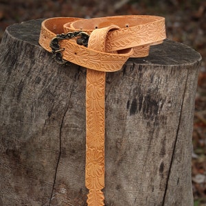 May include: A tan leather belt with a floral design and a brass buckle. The belt is folded and placed on a tree stump.