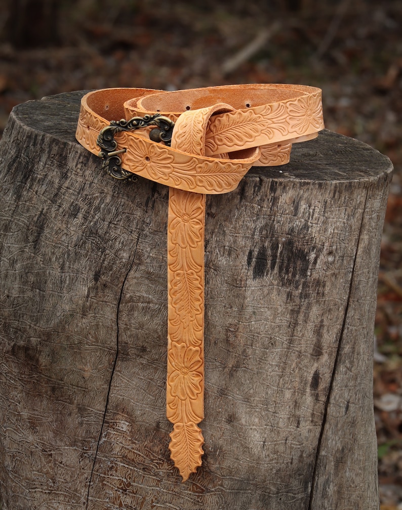 May include: A tan leather belt with a floral design and a brass buckle. The belt is folded and placed on a tree stump.