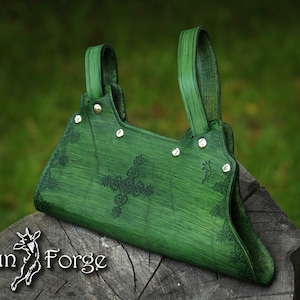May include: A green leather bag with a decorative floral design and silver accents. The bag has a top handle and a flap closure. The bag is sitting on a wooden surface.  Faun Forge.