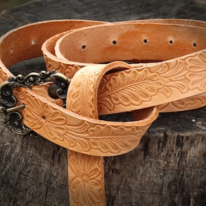 May include: A tan leather belt with a floral design and a brass buckle. The belt is folded and lying on a wooden surface.