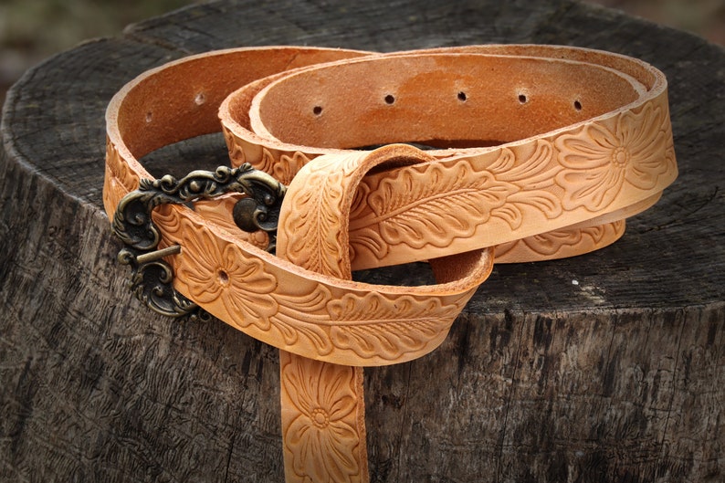May include: A tan leather belt with a floral design and a brass buckle. The belt is folded and lying on a wooden surface.