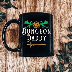 May include: Black ceramic mug with a green and gold design. The mug features two green dragons flanking a gold dice with the text "Dungeon Daddy" in gold and white lettering.