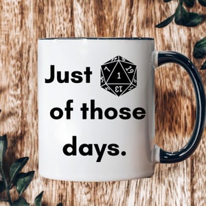 Peut inclure: Une tasse en céramique blanche avec un bord et une poignée noirs. La tasse présente un motif noir d'un dé à vingt faces avec le numéro un affiché. Le texte "Just of those days." est imprimé en noir sous le dé.