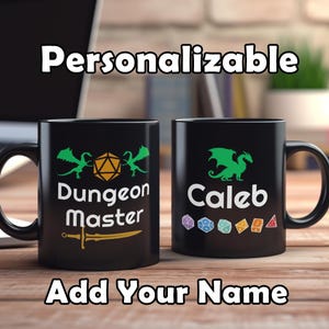 Könnte beinhalten: Zwei schwarze Kaffeetassen mit weißem Design. Die Tasse links hat einen grünen Drachen, einen Würfel und den Text "Dungeon Master". Die Tasse rechts hat einen grünen Drachen und den Text "Caleb".