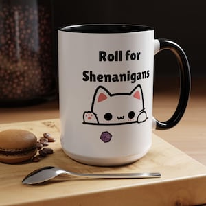 Puede incluir: Taza de cerámica blanca con borde y asa negros. La taza tiene un gato blanco asomándose por la parte superior con un dado morado delante. El texto de la taza dice "Roll for Shenanigans".