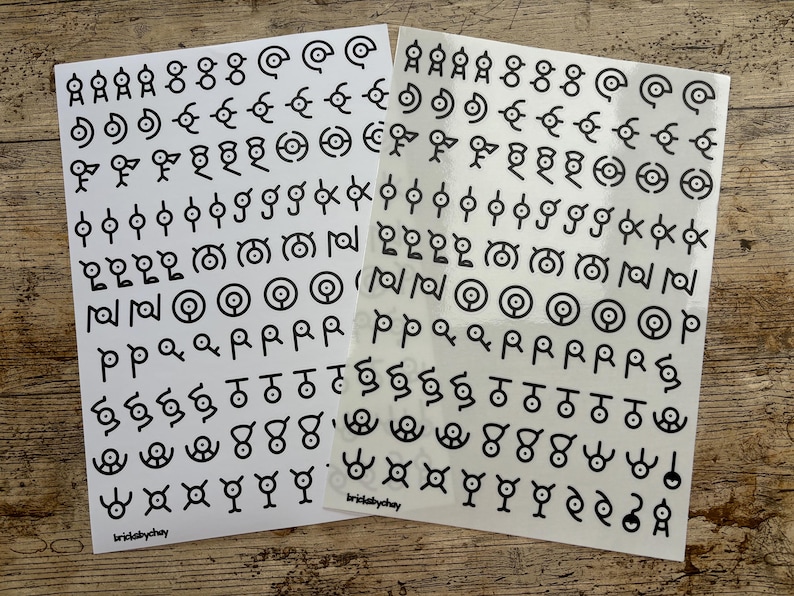 May include: Two sheets of white sticker paper with black symbols. The symbols are various shapes and designs, including circles, squares, and triangles. The text "brickabychay" is printed on each sheet.