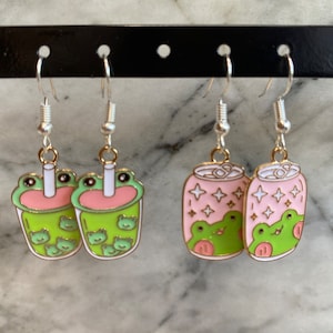 May include: A pair of dangle earrings featuring a green and pink boba tea charm with frog faces and a pair of dangle earrings featuring a green and pink soda can charm with frog faces.