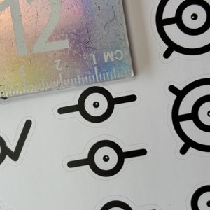 May include: A sheet of white paper with black and white stickers of a cartoon eye with a circle around it. Some of the stickers have a horizontal line across the eye.