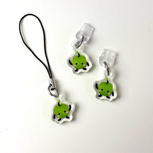 Op de afbeelding: Drie heldere acryl bedels met een groen appelontwerp. Eén bedel is bevestigd aan een zwart koord. De appel heeft een lachend gezicht en zwarte contouren. Twee bedels hebben doorzichtige plastic clips.