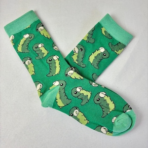 以下が含まれることがあります： 漫画風の緑と薄緑の生き物が繰り返されるパターンの緑色の靴下。 靴下は薄緑色の袖口とつま先が付いています。 靴下は柔らかい素材で作られています。