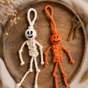 May include: Two macrame keychains shaped like figures, one white and one orange. Each features a round wooden bead head with a hand-painted black jack-o'-lantern face and a knotted cord body with arms and legs. A metal key ring is attached.