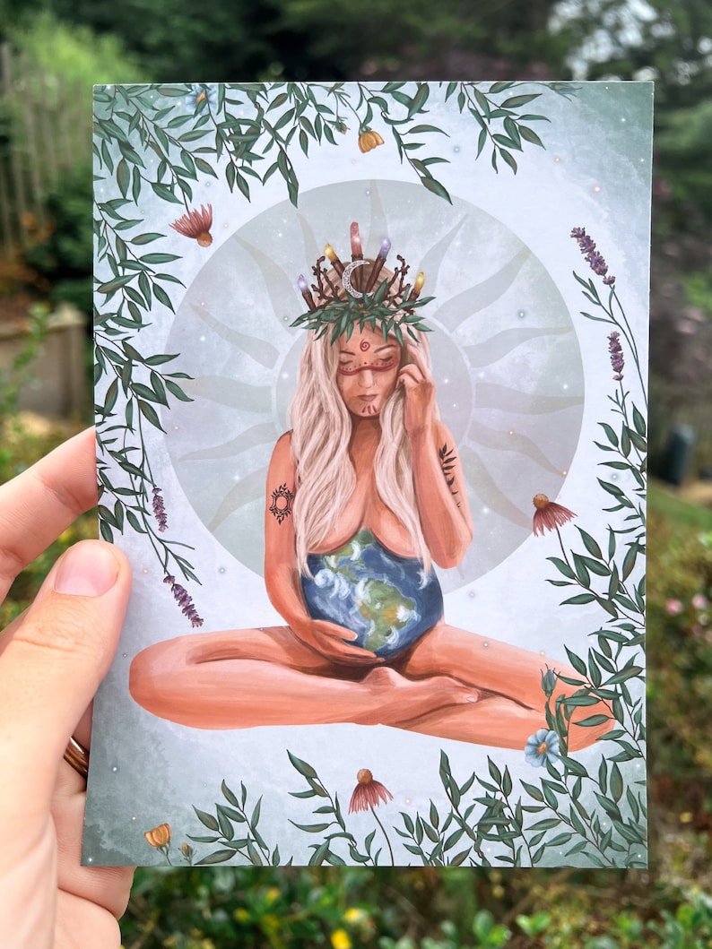 May include: A woman with a crown of leaves and candles sits with her legs crossed, holding a globe in her lap. She has a painted face and tattoos on her arms. The background is a soft blue with green leaves and flowers.