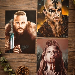 May include: Three digital art prints featuring characters from the TV show Vikings. The first print shows a man with a beard and a sword. The second print shows a man with a beard and a rune on his forehead. The third print shows a woman with red hair and blood on her hands.